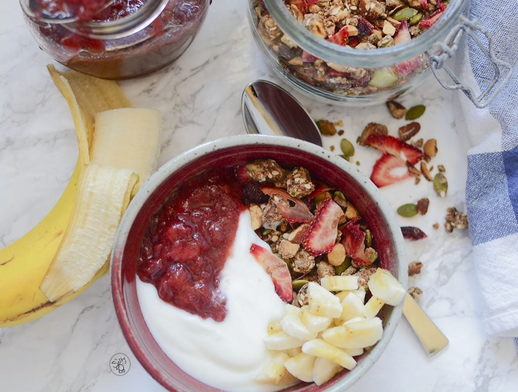 A granola bowl with yogurt, jam and bananas on top. A half of a banana lays on the table to the left, and above it are the jars of jam and granola.