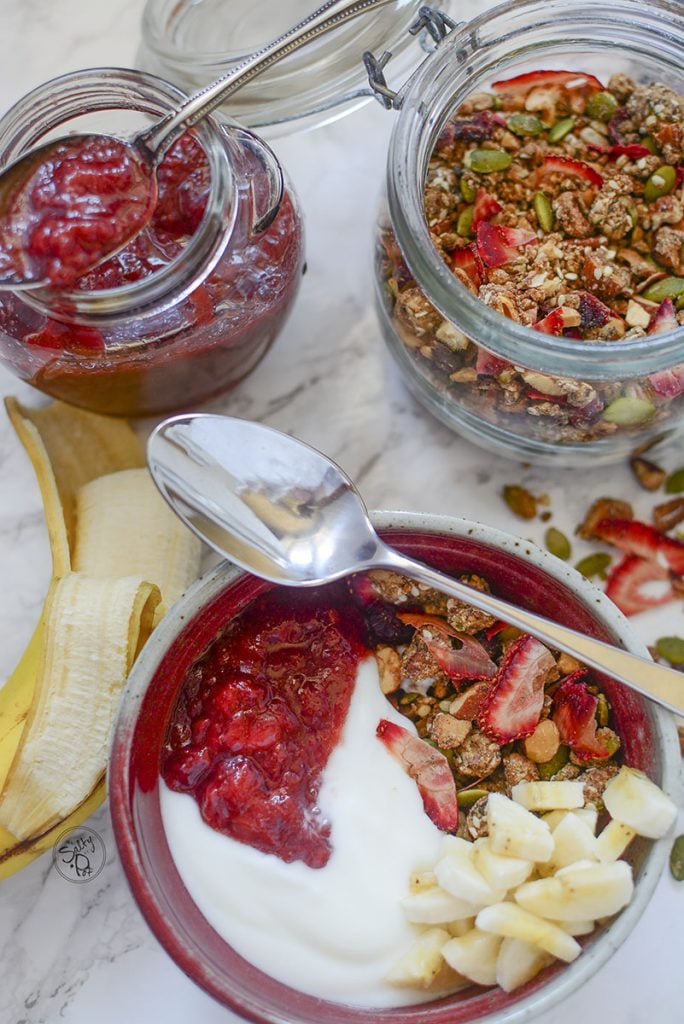 A top down photo of the breakfast table. Granola bowl on the bottom, a spoon resting on top of the bowl. The jam jar and granola jar sit to the top of the bowl with half a banana on the left. 