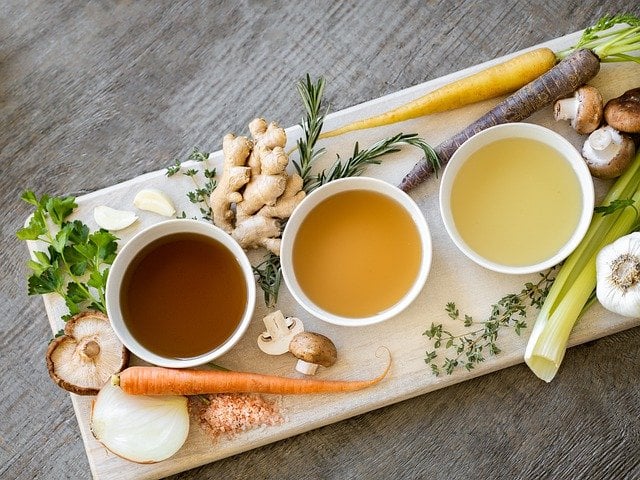 Three types of bone broth sitting on a wood board with a variety of vegetables and aromatics sitting next to them.