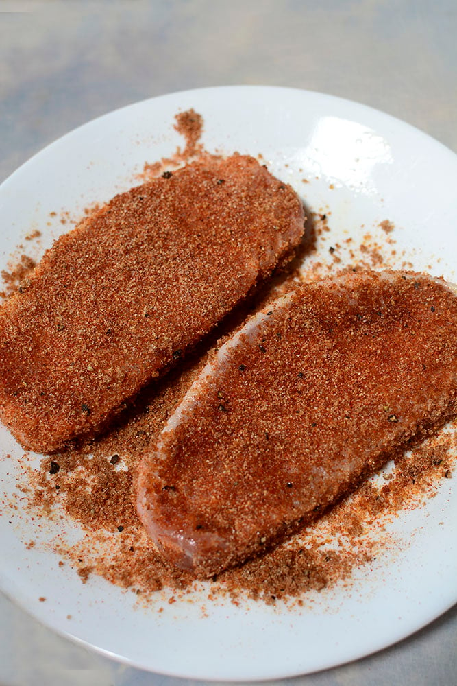 Two pork chops with seasoning on them, on a white plate.