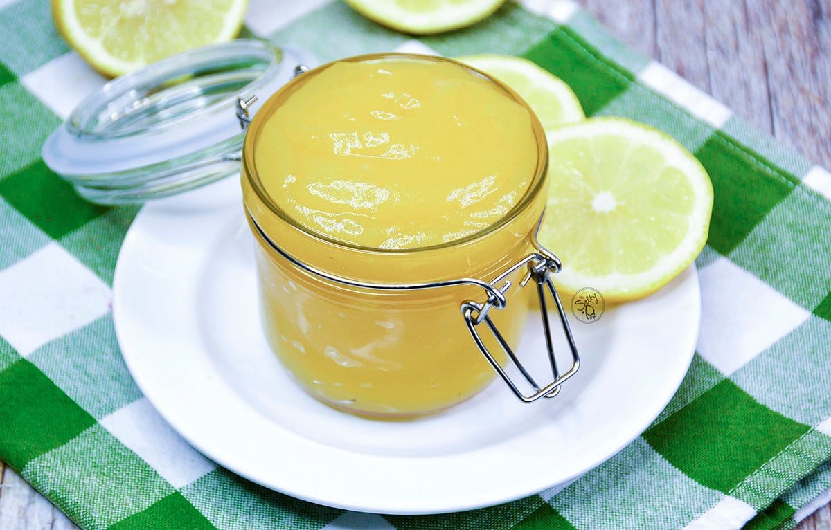 Lemon curd in a pretty jar sitting on a white plate with lemon slices around it.