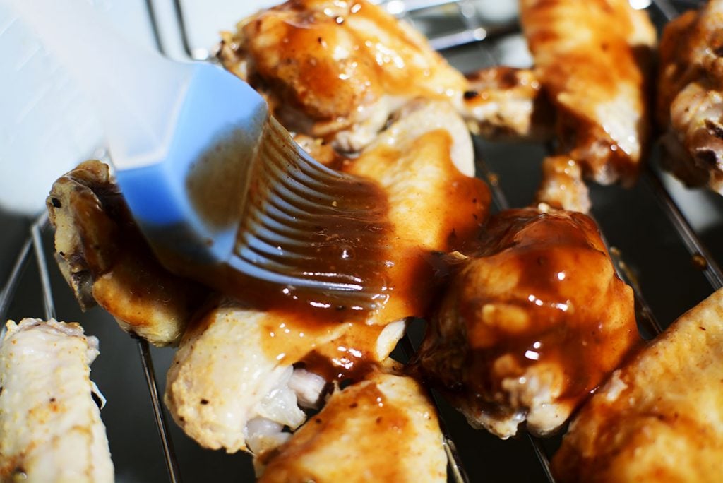 A blue silicone basting brush is spreading bbq sauce on top of the wings.