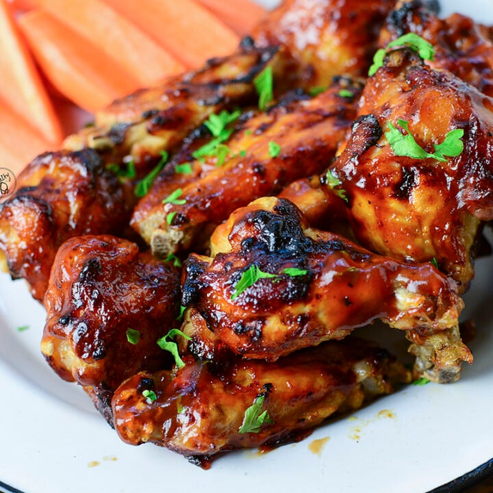 A dozen bbq chicken wings sitting on a white plate with cut carrot sticks in the background.