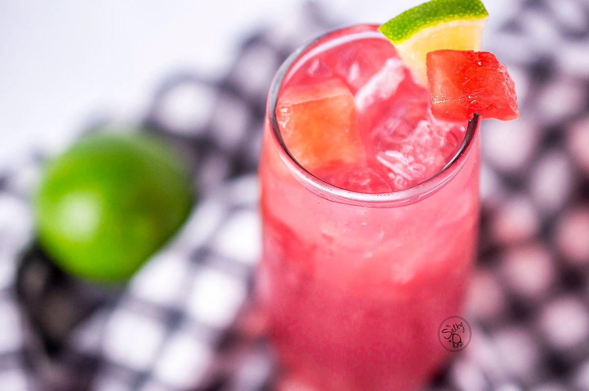 Watermelon punch in a tall glass with a green lime in the background. The glass is resting on a checkered napkin.