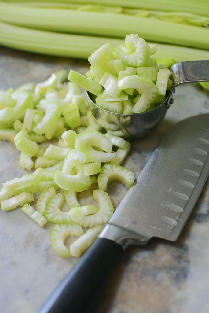 A bunch of cut celery in half moon shapes are resting on the cutting board, and in a measuring cup. A knife is sitting beside the celery. 