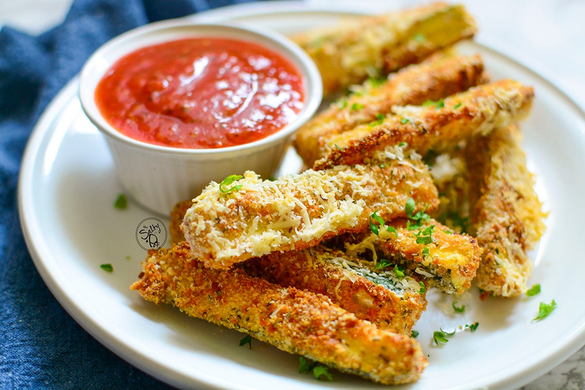 Zucchini sticks on a white plate with marinara sauce in the background.