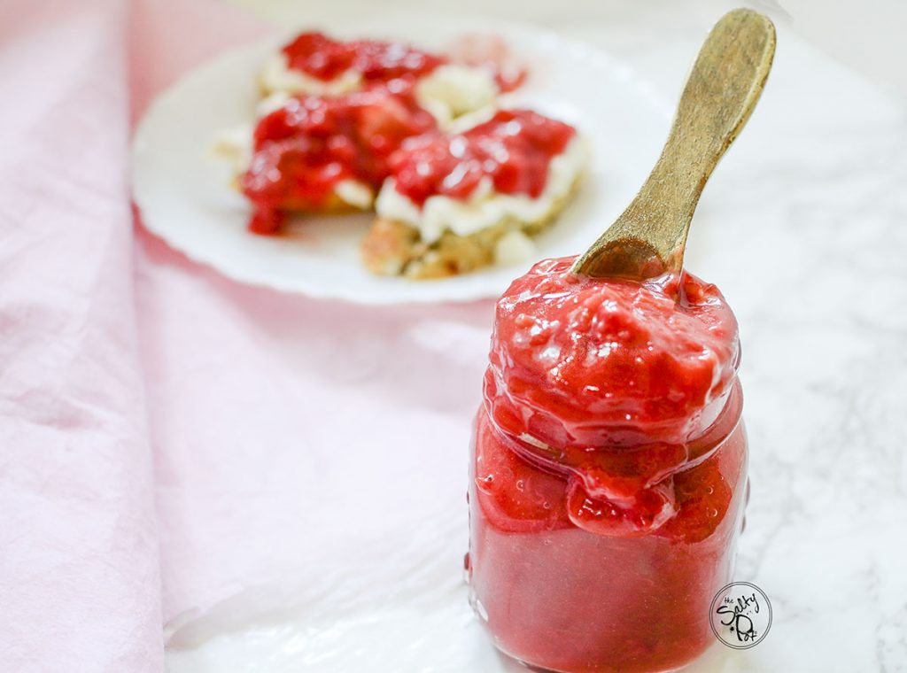 Strawberry Rhubarb sauce overflowing in a jar with a spoon sticking out the top of it. In the background is a plate with toast on it.
