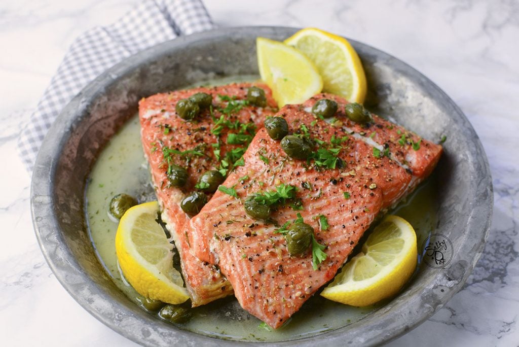 Air fryer salmon sitting on a plate with lemon wedges sitting around it, along with capers on top.