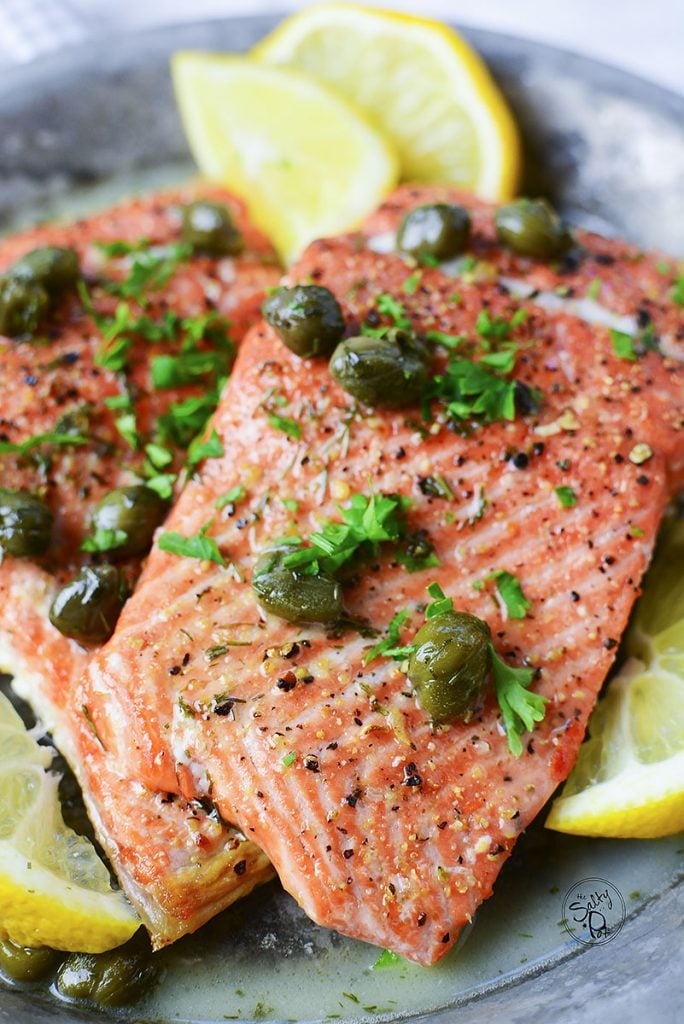 A close up photo of a cooked salmon filet with lemon wedges and green capers on top.