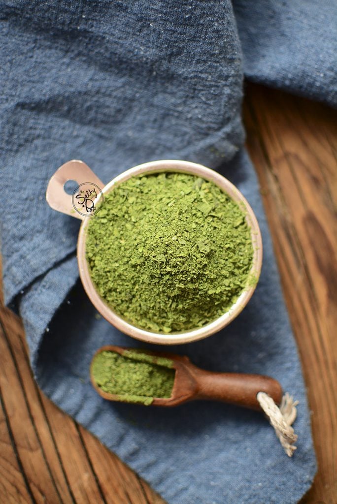 Spinach powder in a copper tin with a wooden spoon beside it on a blue napkin.