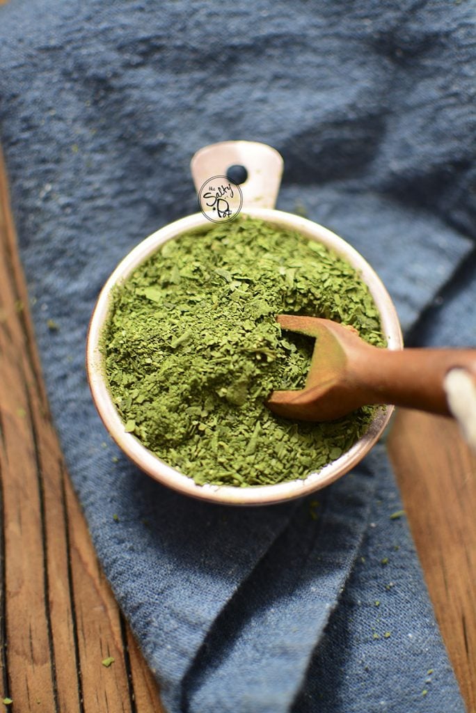 Spinach powder in a cup with a wooden spoon sticking out to the right.