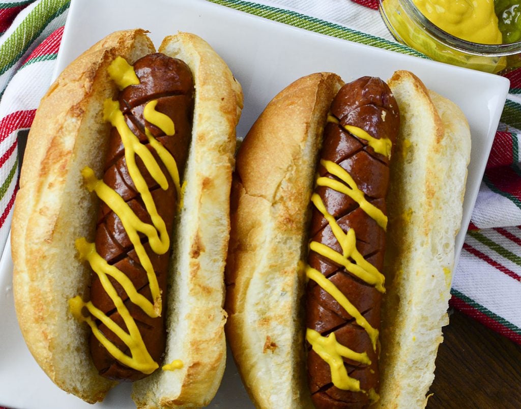 A top down photo of two air fried hot dogs in buns, sitting on a white plate with a colorful napkin in the background.