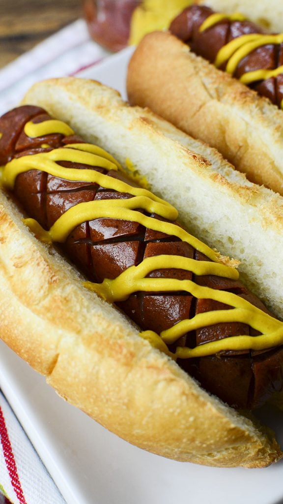 It's easy to learn how to air fry hot dogs. This is a photo of an air fried hot dog sitting in a toasted bun with mustard over the top, sitting on a white plate.
