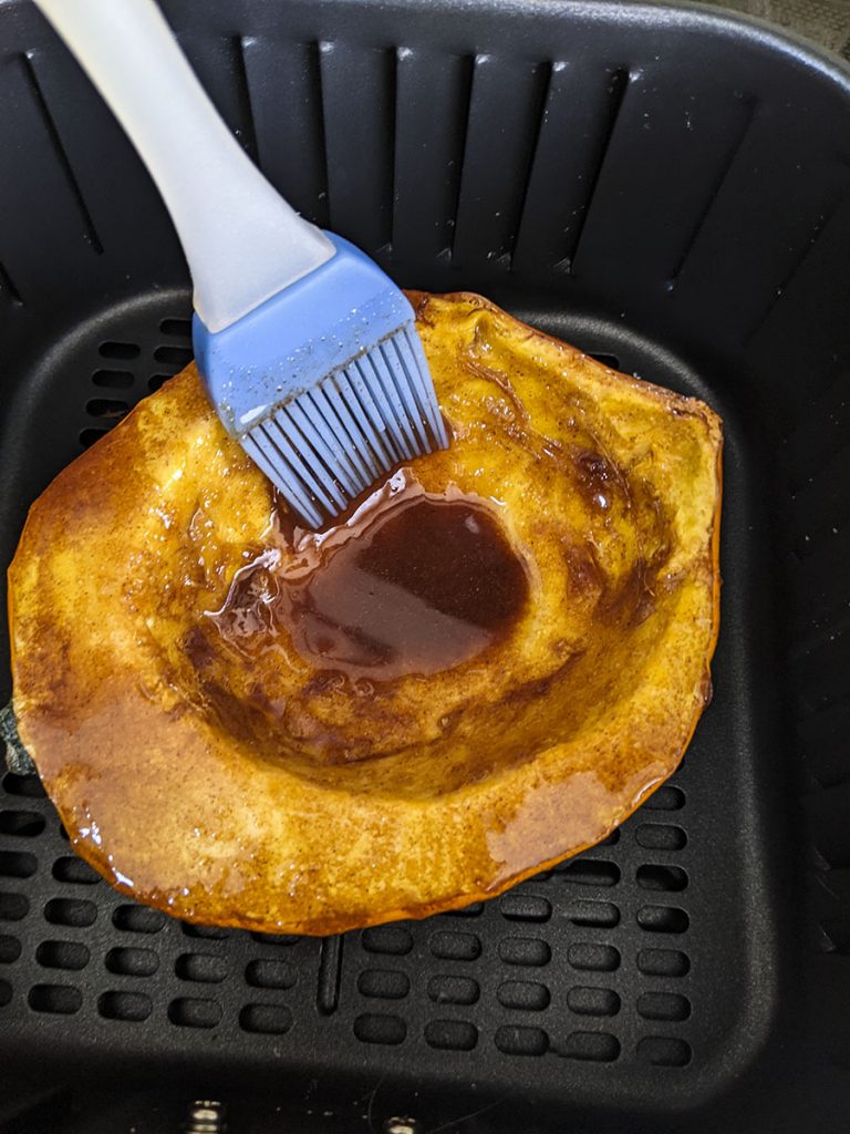 The one half of an acorn squash inside the basket of the air fryer. The buttery cinnamon topping is being basted on top of the vegetable. 