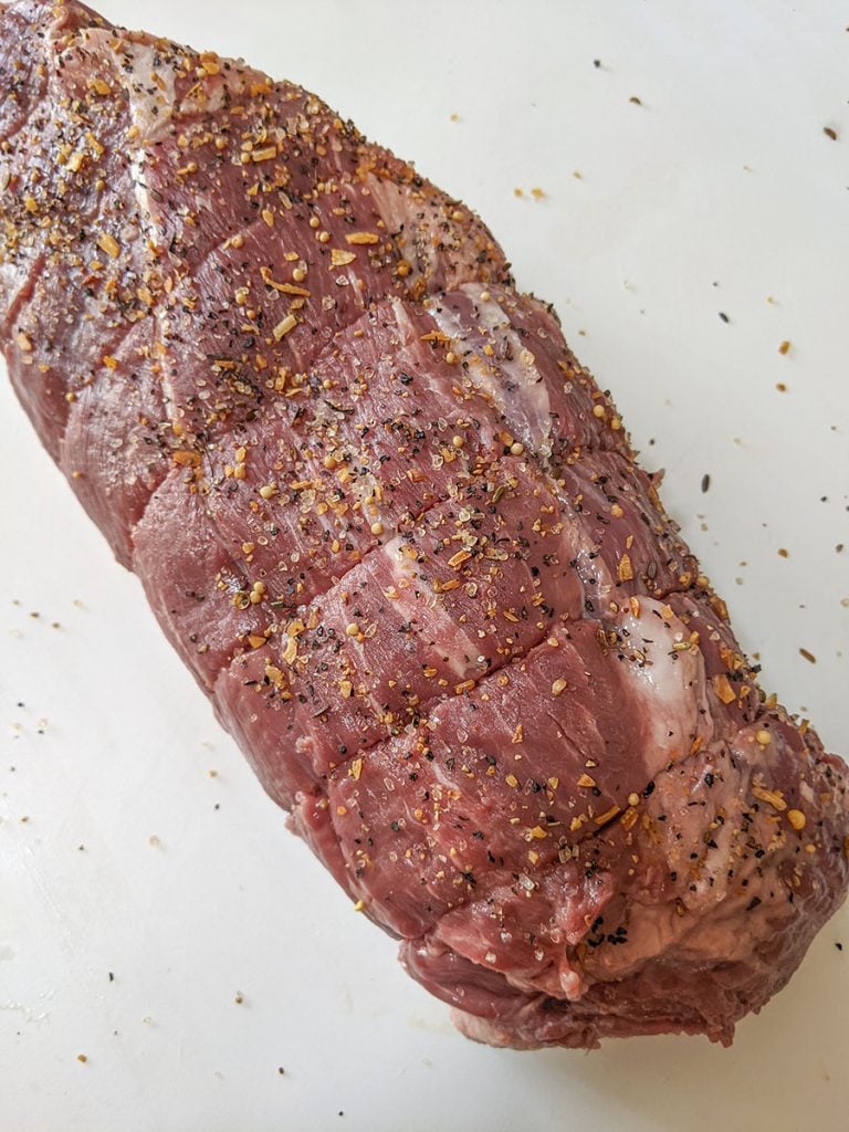 The roast, sitting on a white cutting plastic cutting board, covered with the seasoning spice.