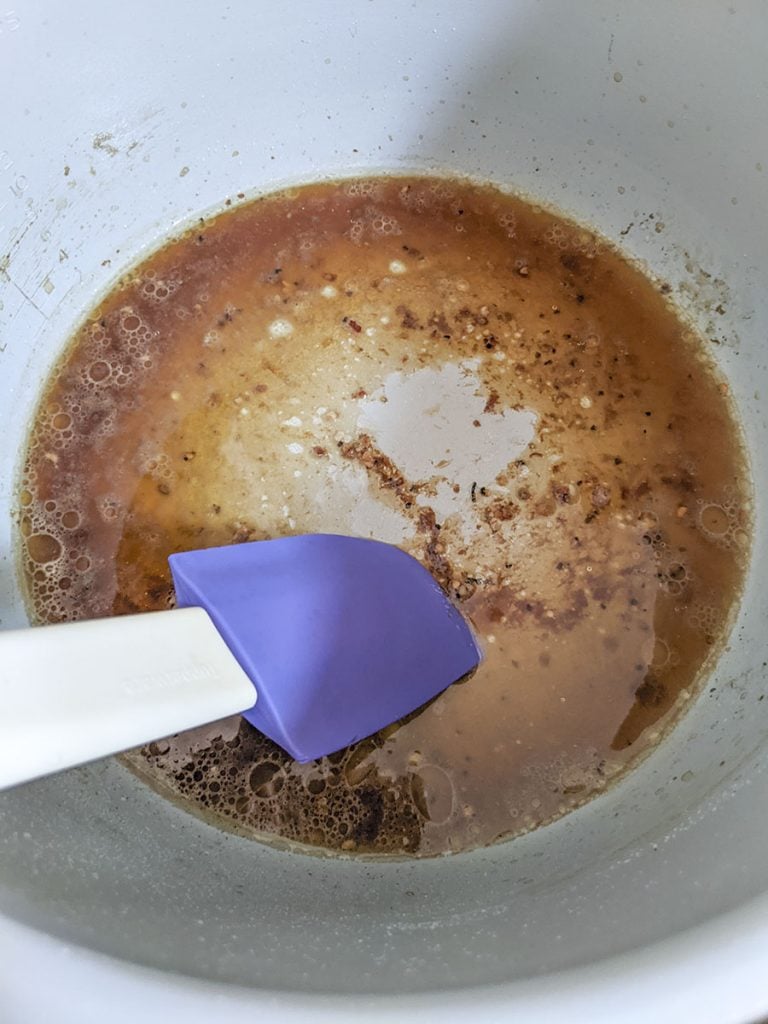 A blue spatula is lifting the fond off the bottom of the cooking pot after pouring a little bit of broth into it. 