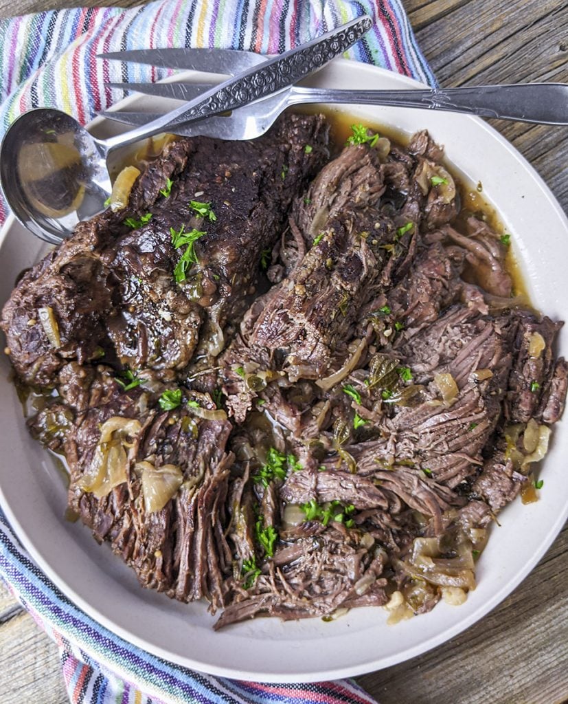 A cooked beef roast sitting on a white plate with a gravy spoon and a large serving fork laying at the top.