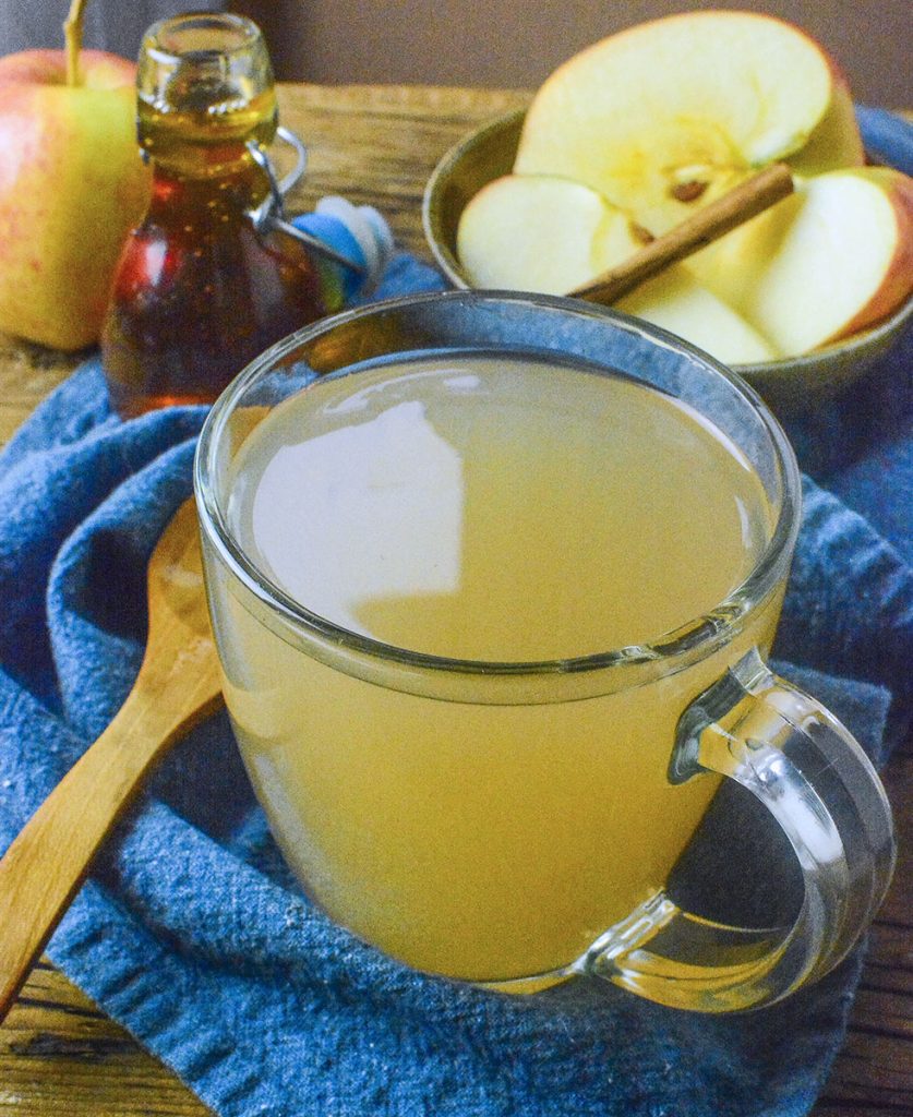 A clear mug holds apple cider with a wooden spoon on the left and apples and honey in the background.