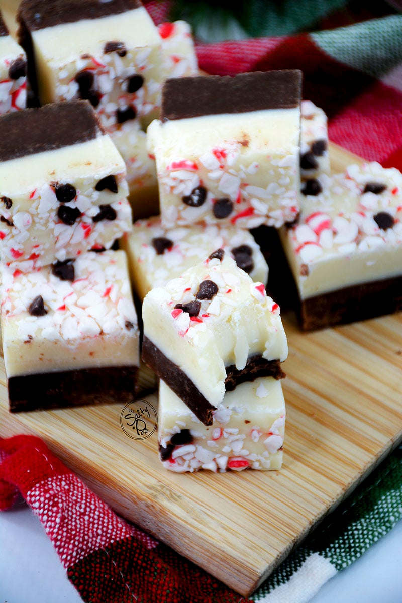 Pieces of fudge sitting on a wooden board. Fudgy brown bottom with a white vanilla top. Covered in red crushed candy canes and mini chocolate chips.