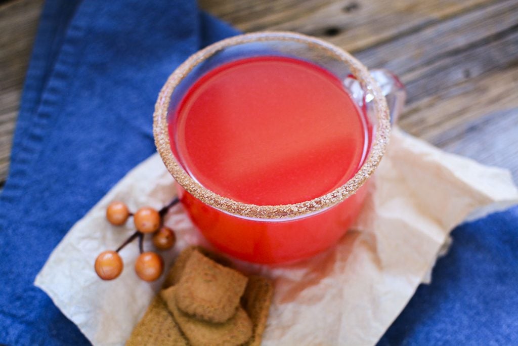 Red hot apple cider in a clear glass jar with a cinnamon sugar rim. A few cookies are sitting at the base fo the glass.