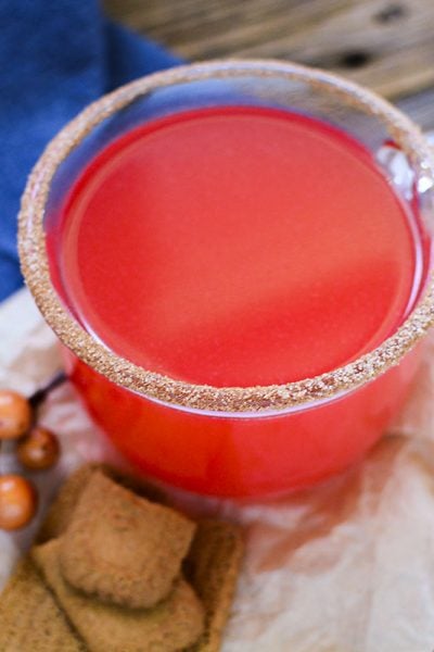 Red hot apple cider in a clear glass jar with a cinnamon sugar rim. A few cookies are sitting at the base fo the glass.