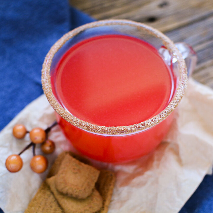Red hot apple cider in a clear glass jar with a cinnamon sugar rim. A few cookies are sitting at the base fo the glass.