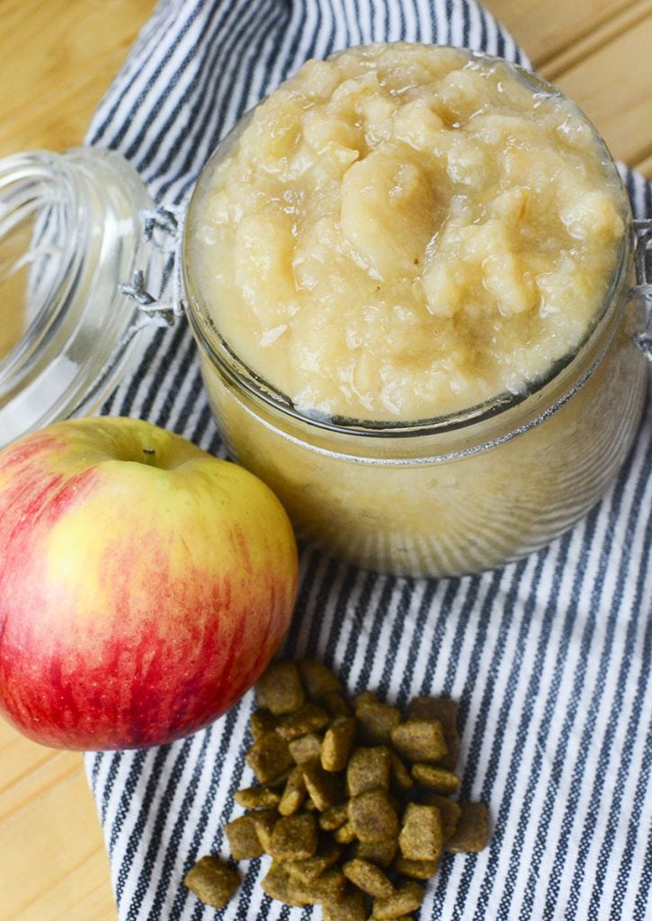 2 cups of applesauce that's made for dogs sitting in a glass jar. An apple and some dog kibble rests beside it.