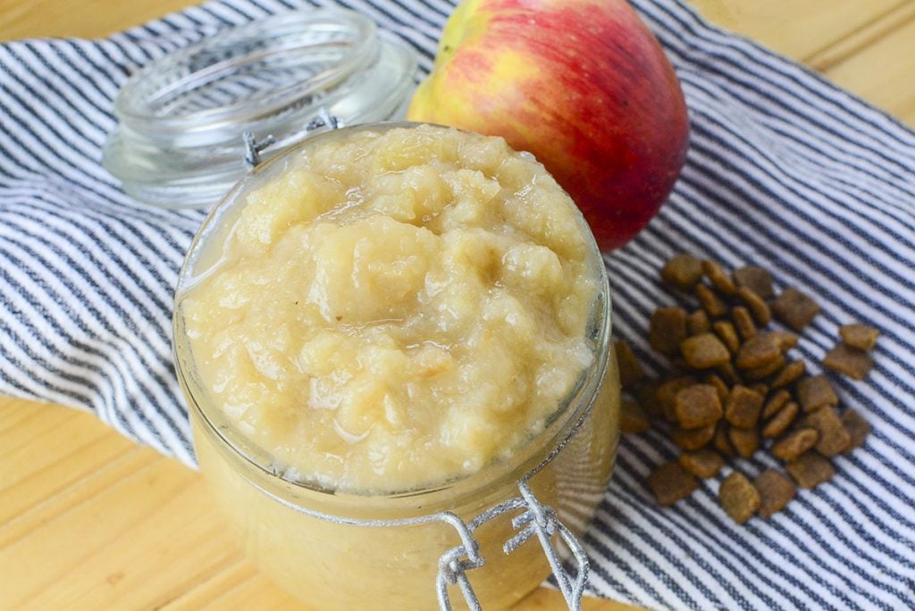 Finished applesauce in a glass container. On the right is a small bundle of kibble with an apple sitting at the top of the photo.