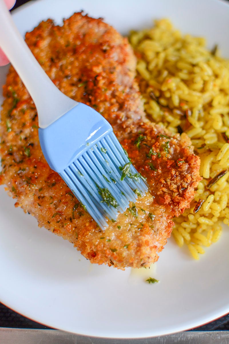 A blue silicone basting brush spreads butter with parsley over the top of the pork steak.