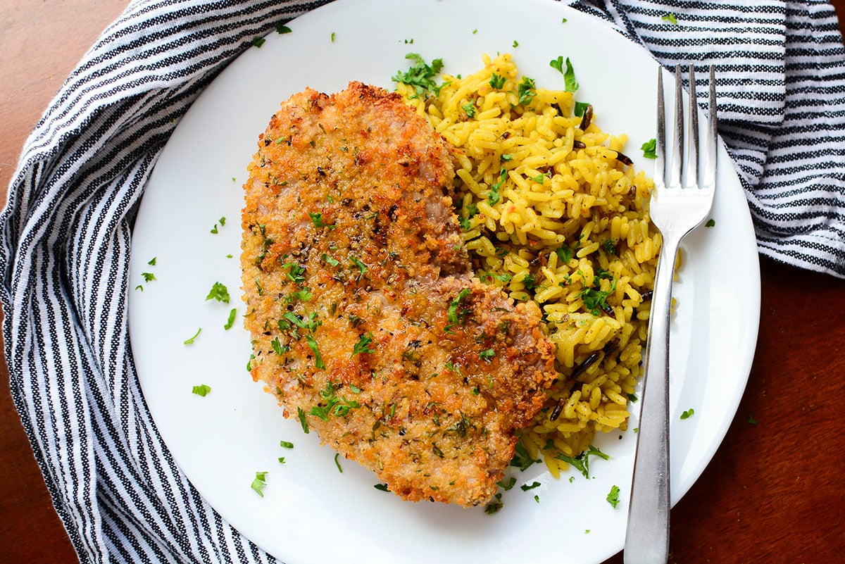 Air fryer pork steak next to a side of rice on a white plate. There is a fork on the right side of the plate.