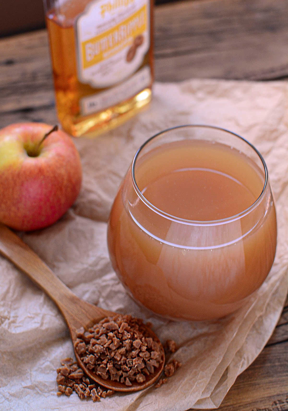 A glass of toffee apple cider sits on a table with an apple on the left, toffee bits down in the front and a small bottle of liquor in the background.