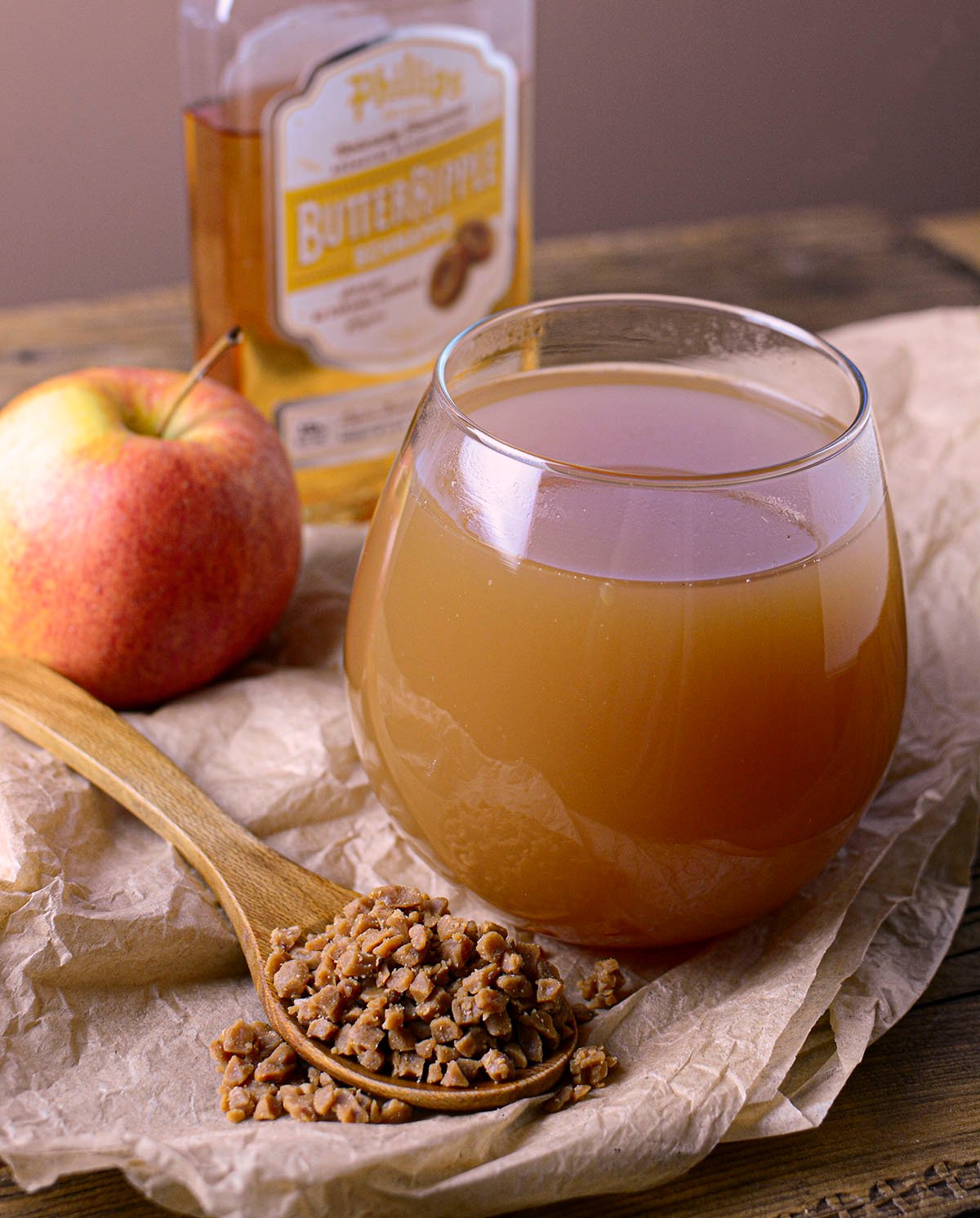 Warm and comfortaing, toffee apple cider sits on a wooden board with toffee bits in a wooden spoon in front of it.