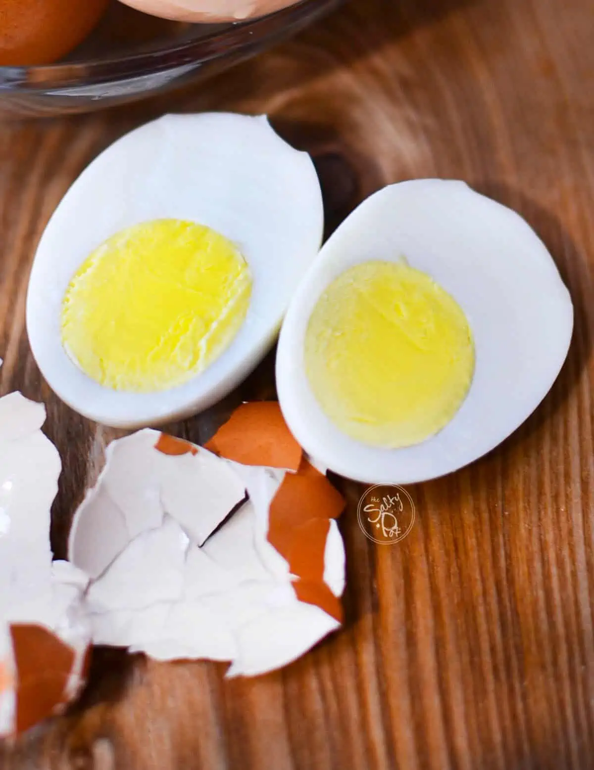 Two perfectly cooked hard boiled eggs on a wooden cutting board with the shell sitting beside it.