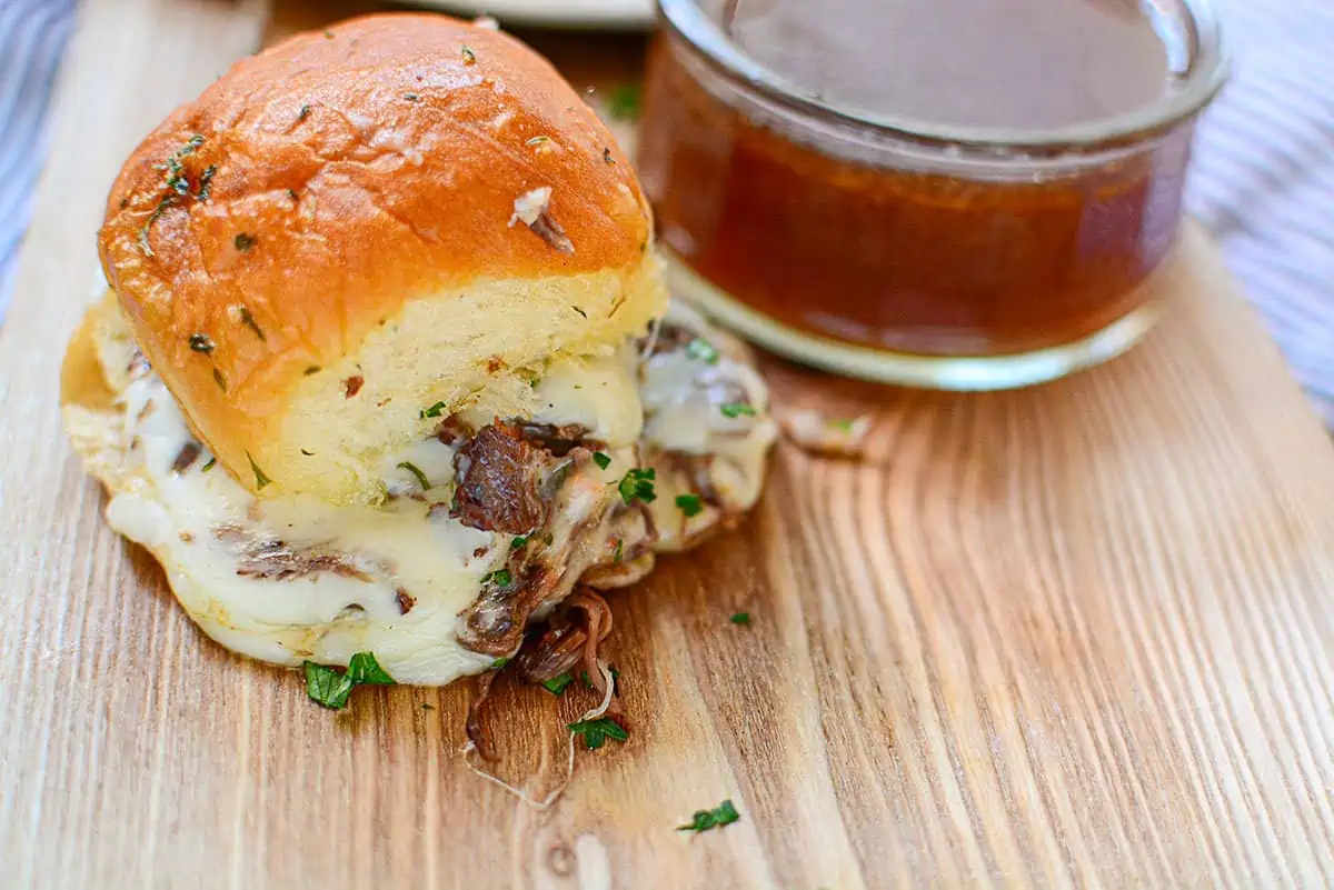 Roast beef slider with au jus dipping sauce on a wooden cutting board.