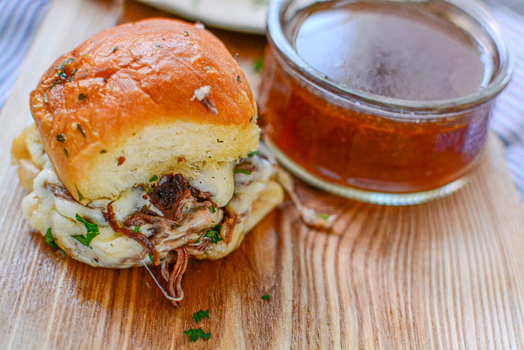 The roast beef slider sits on a wooden board next to a bowl of au jus.