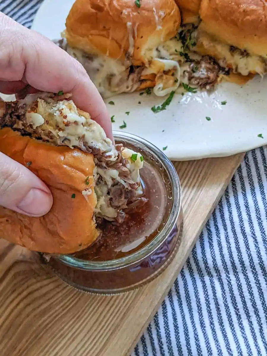 A beef slider dipped into au just sauce with other sliders in the background. 