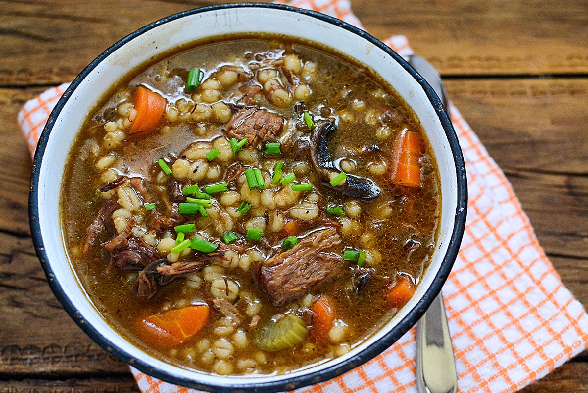 Beef stew with barley in a white enamel bowl on a wood background.
