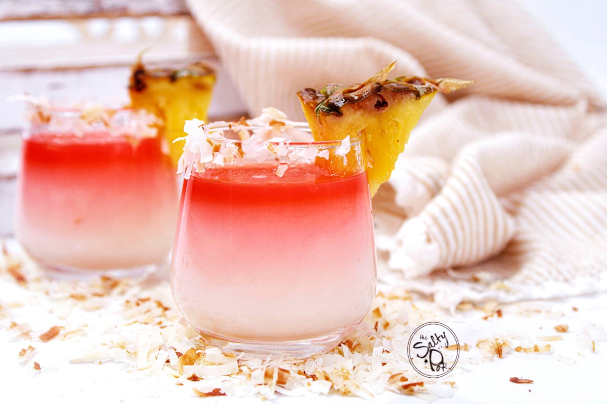 Two filled shot glasses surrounded by coconut flakes and a beach blanket.
