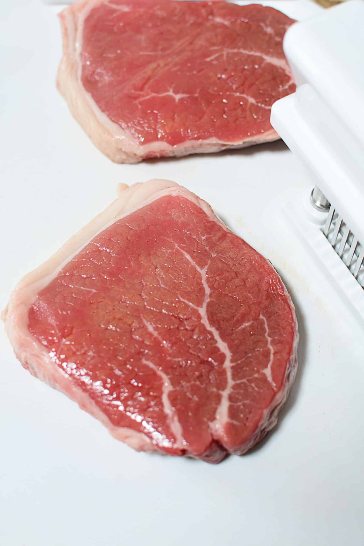 Two tough steaks with the tenderizer next to it on the right.