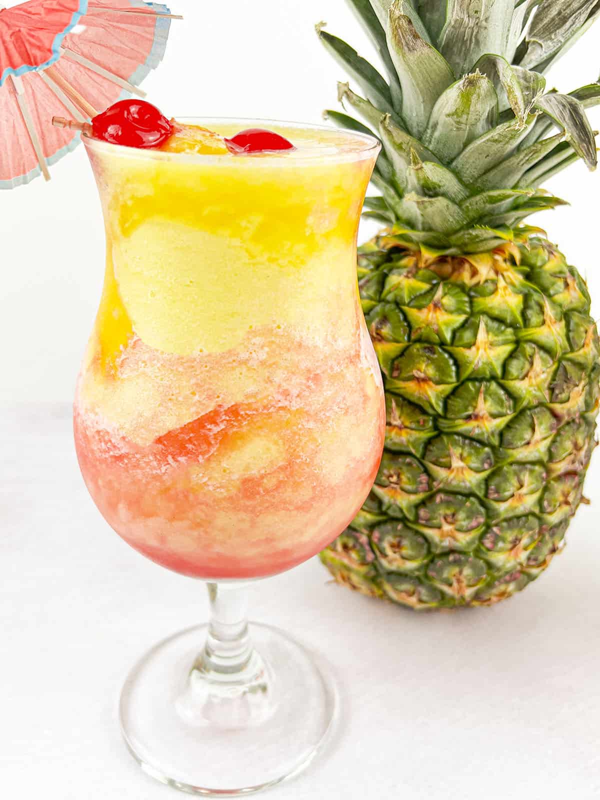 The fruity drink with cherries and a paper umbrella in the glass next to a pineapple.