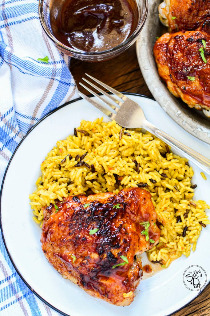 BBQ chicken thigh on a plate with rice served next to it. At the top is a small bowl of BBQ sauce for dipping.