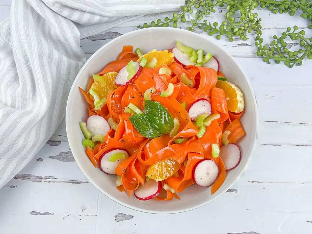 Orange shaved ribbons of carrots with radish rounds, orange slices and celery on a white wooden backdrop.