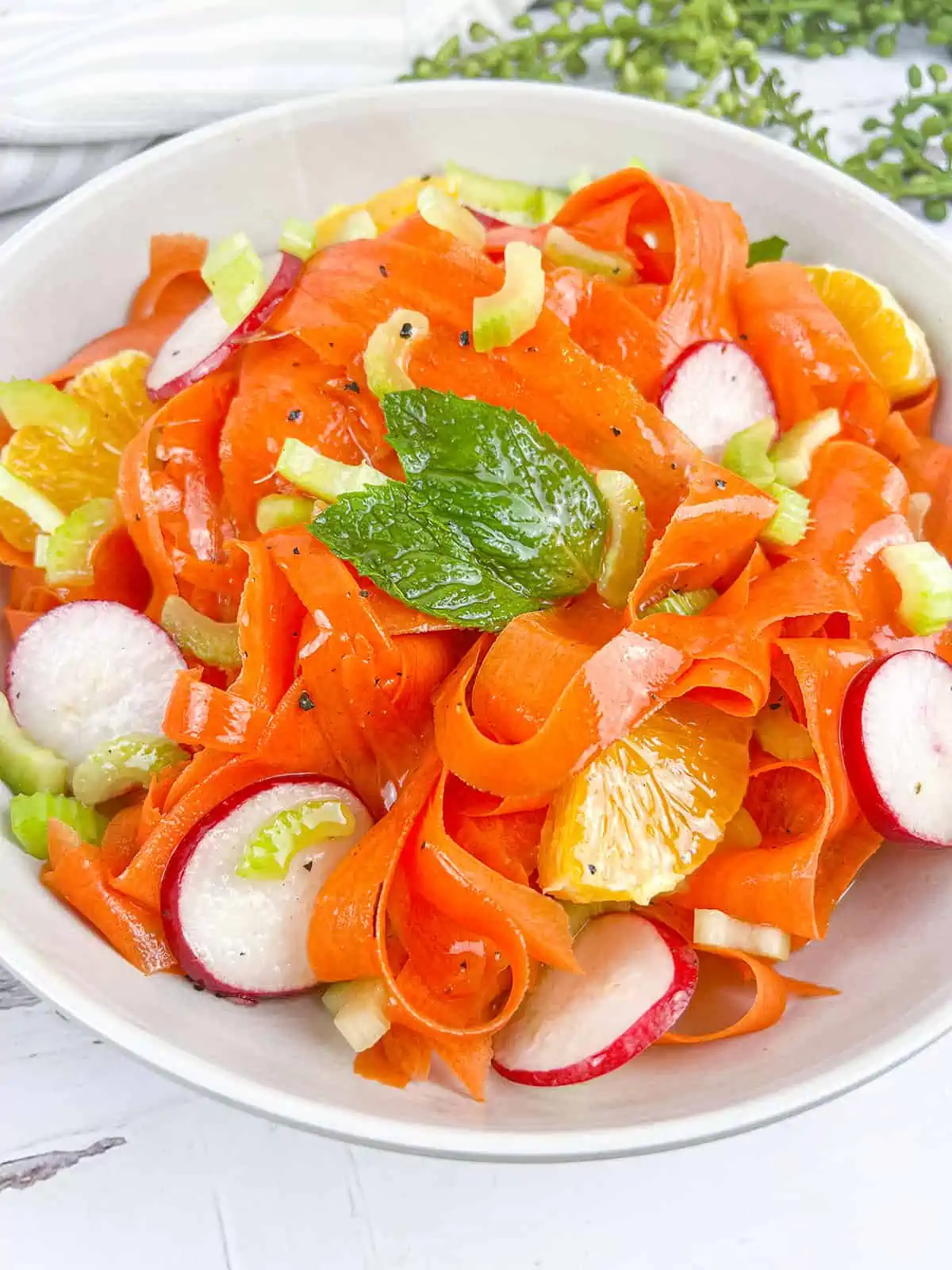 Shaved carrots, radishes, orange segments, celery and mint leaves all in a white bowl with dressing over top.