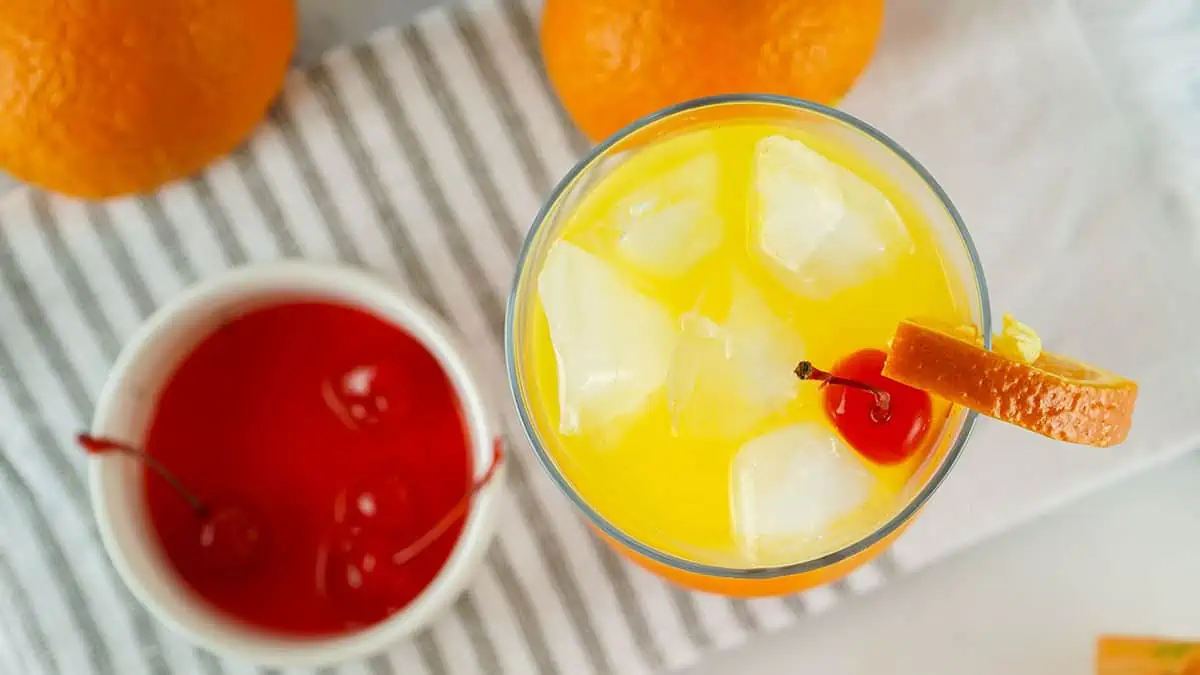 Photographed from above, the cocktail is on the right and a small bowl of cherries is on the left. Oranges are at the top.