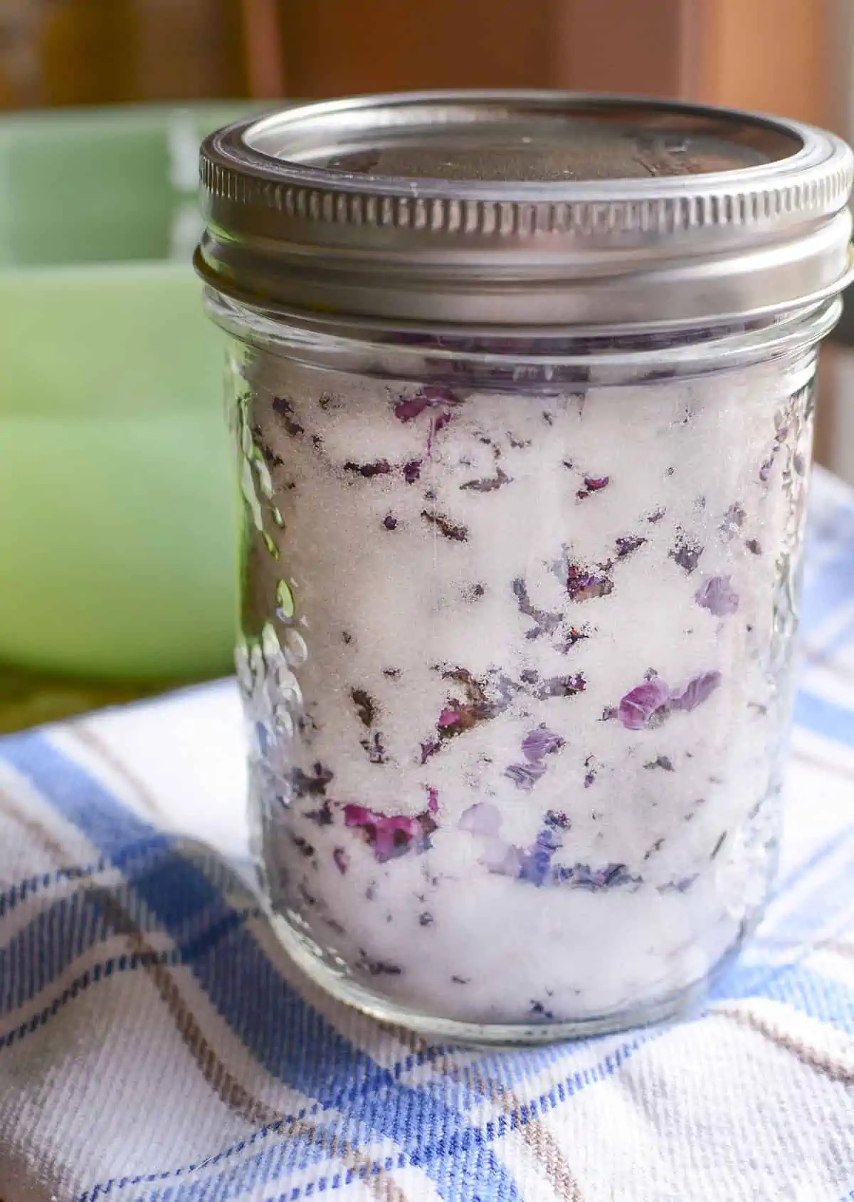 Lilac sugar in a mason jar with a lid on it. The jar is resting on a blue and white checked cloth.