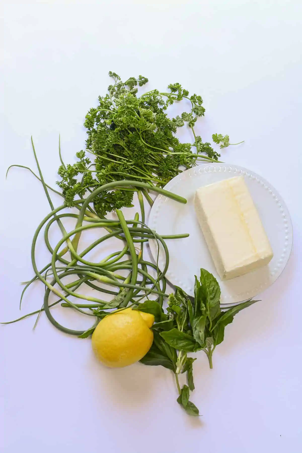 The ingredients needed to make garlic scape butter. Garlic scapes, butter, lemon, parsley and basil.