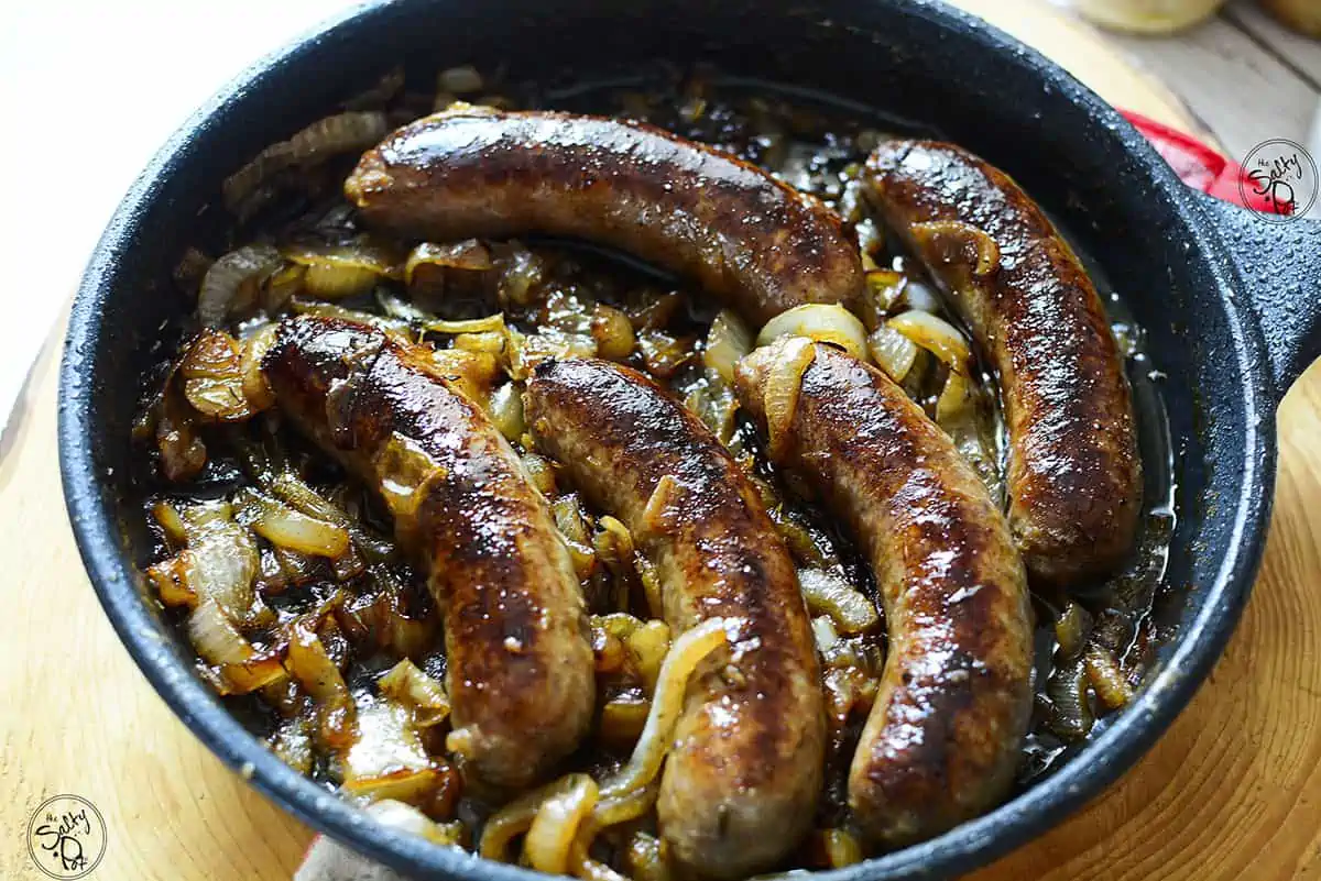 Five brat sausages with caramelized onions sit in a cast iron skillet on a wooden board.
