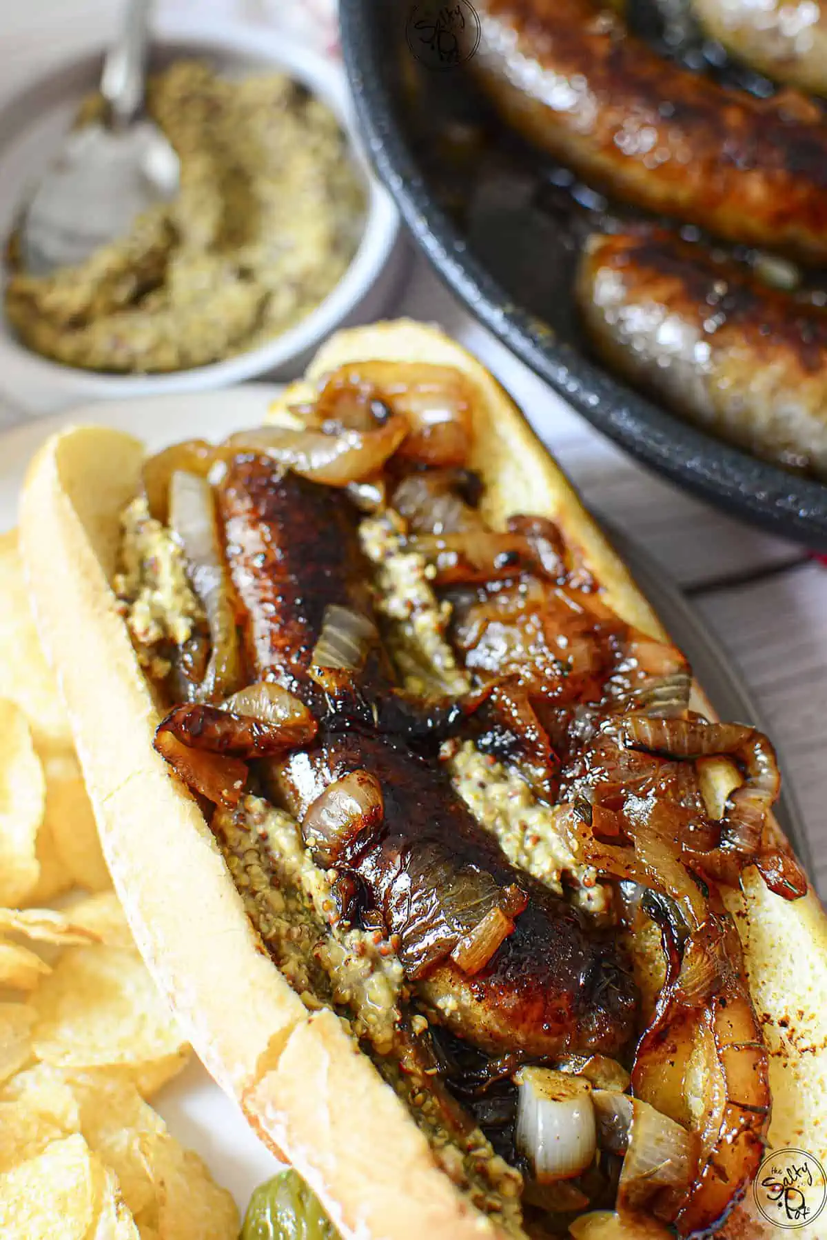 A brat in a bun with onions and condiments. In the background are more brats in a pan.