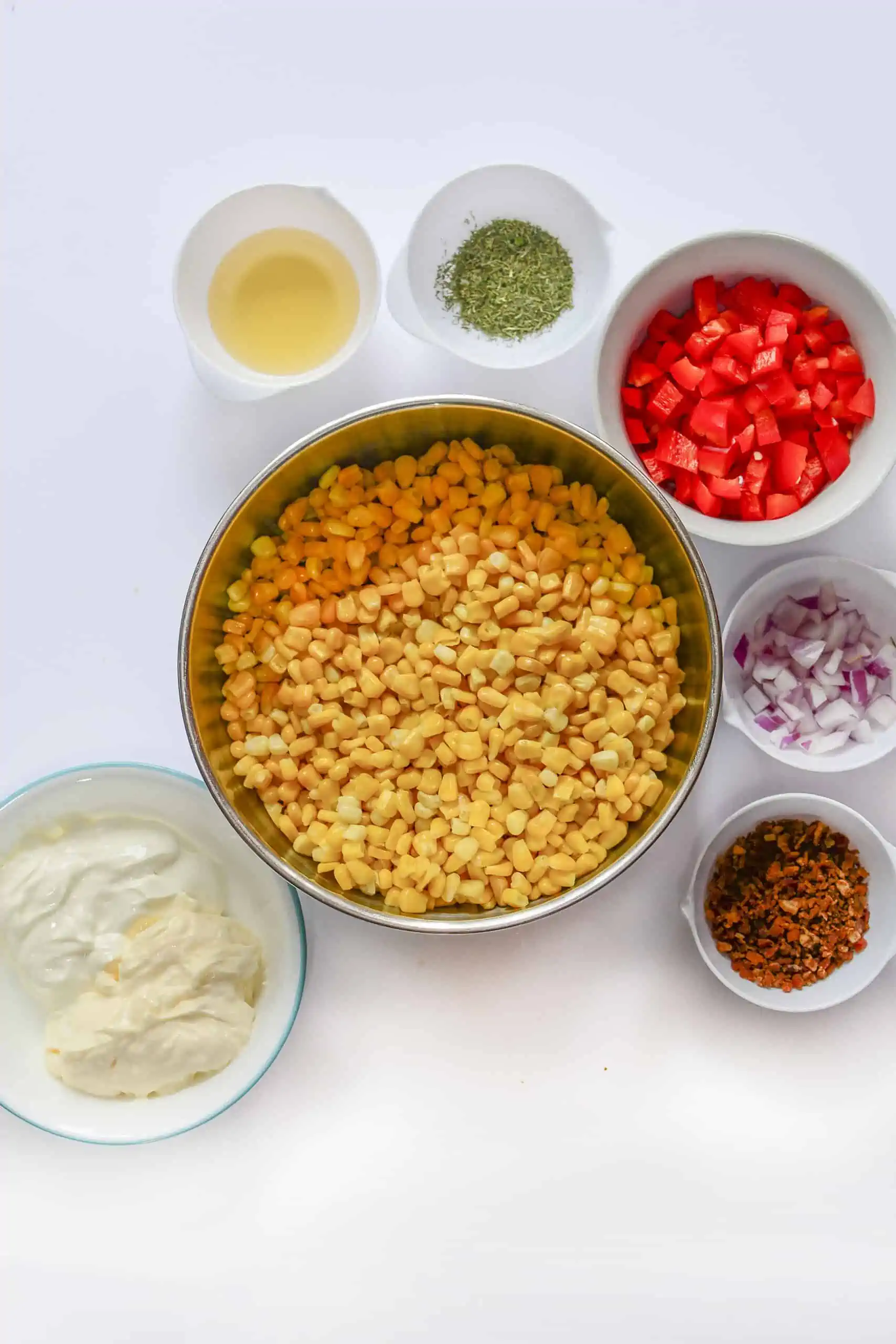 The ingredients needed for this salad - corn in the center. Above is vinegar, dill, tomatoes, red onion, bacon, mayonaise and sour cream in a bowl at the bottom.
