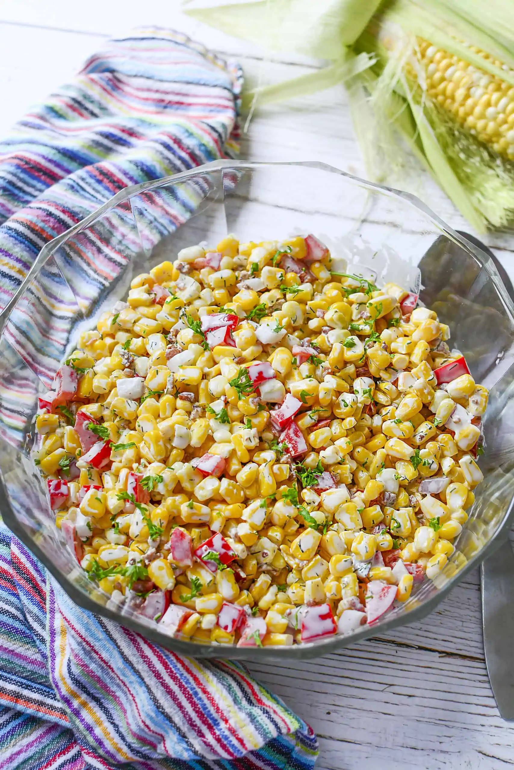 Corn salad in a glass bowl with a silver serving spoon next to it. On the left is a colorful striped napkin.